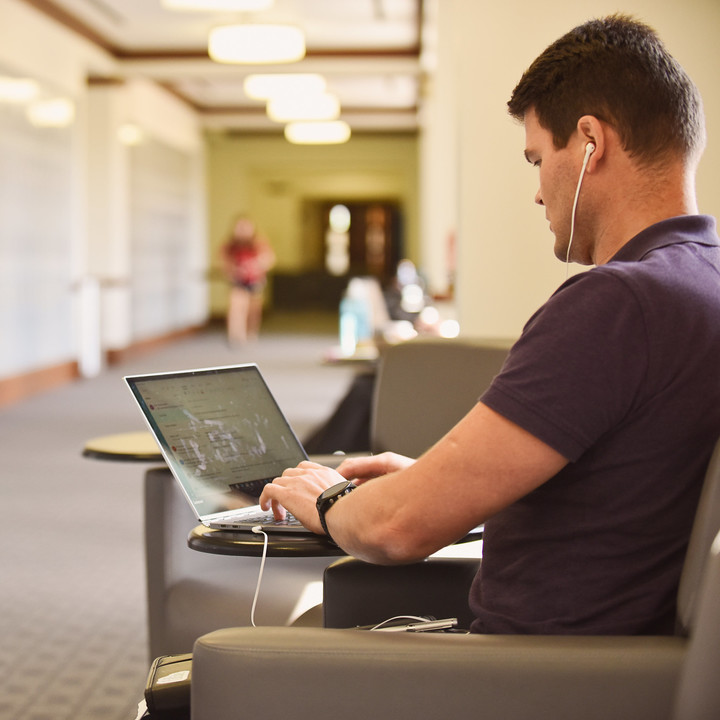 Male MBA student in purple collared shirt with headphones on computer in hallway